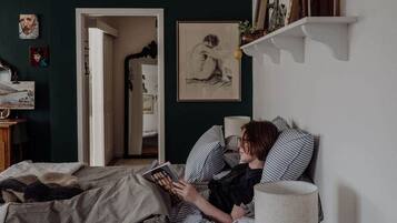 Eine Person liest ein Buch in einem Zimmer mit einem Bett, einer Lampe und einem gerahmten Bild an der Wand.