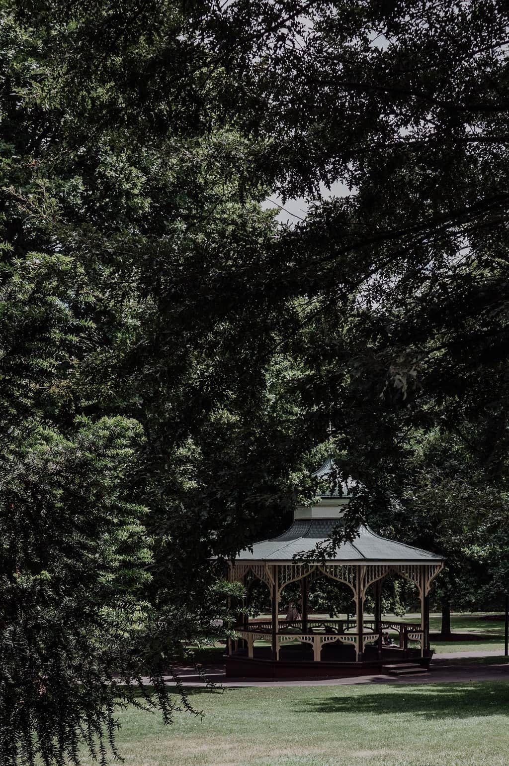 Um coreto com cobertura e laterais abertas, cercado por gramado e árvores.