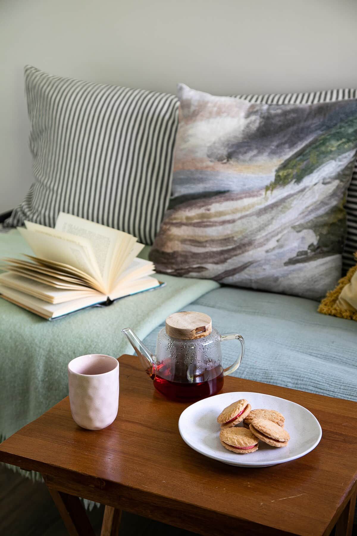 A cozy reading nook with a bed, pillows, an open book, a mug, a teapot, and biscuits on a small table.