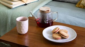 Een gezellig hoekje om te lezen met een bed, kussens, een open boek, een mok, een theepot en koekjes op een klein tafeltje.