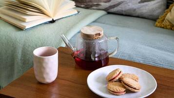 A cozy reading nook with a bed, pillows, an open book, a mug, a teapot, and biscuits on a small table.