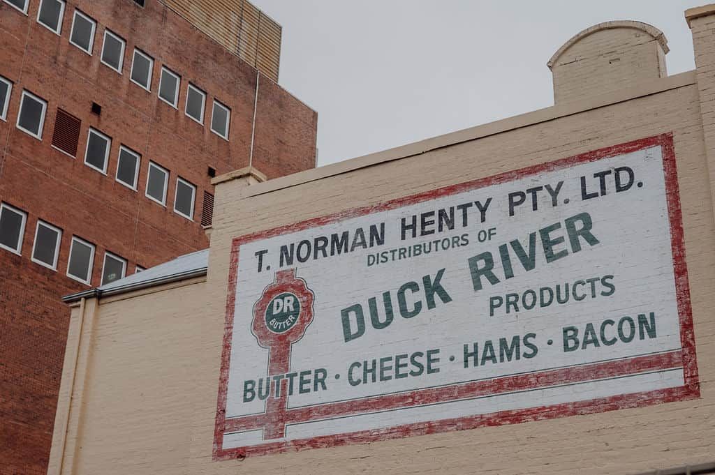 A building with a sign for T. Norman Henty Pty. Ltd., distributors of Duck River products like butter, cheese, hams, and bacon.