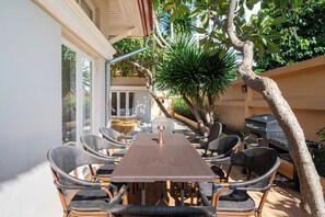 Un espace extérieur aménagé pour les repas, avec une table en bois et des chaises en osier, entouré de verdure et agrémenté d’un barbecue.