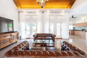 A spacious living room with a leather sofa, a glass coffee table, and a television on a wooden cabinet. - Naka Pool Villa Pratumnak Pattaya (Pattaya City)