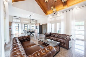 A spacious living room with a leather sofa, a glass coffee table, and a dining area with a chandelier. - Naka Pool Villa Pratumnak Pattaya (Pattaya City)