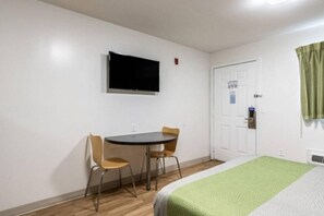 Une petite pièce comprenant un lit, un bureau avec deux chaises, un téléviseur fixé au mur et une porte avec une fenêtre.