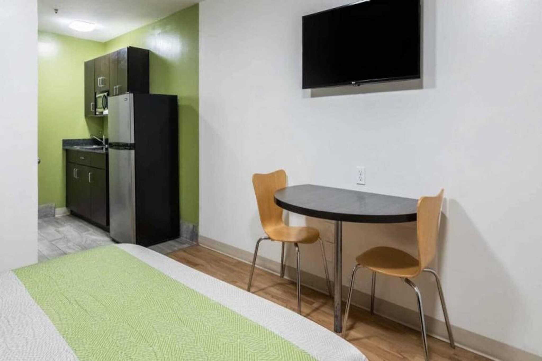 A room with a bed, a small kitchenette, a TV mounted on the wall, and two chairs around a table.
