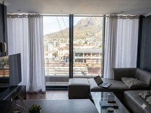 A modern living room with a sofa, a coffee table, and a laptop. There is a large window with curtains, offering a view of a cityscape and mountains. - Serene 1-bedroom Apartment in Central Cape Town (Cape Town)