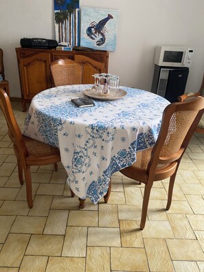 A dining area with a round table covered by a blue and white patterned tablecloth, surrounded by wooden chairs with woven seats. - Vrbo Property (Bains-sur-Oust)
