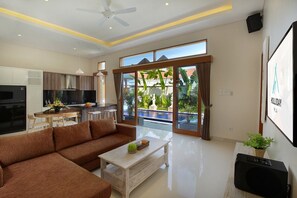 A modern living room with a sofa, coffee table, and a TV. There is a kitchen area with a refrigerator and a dining table. The room has large windows and a view of a swimming pool. - Holl Villa - Family Two Bedroom Villa (Kuta)