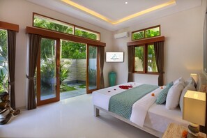 A bedroom with a bed, curtains, a TV, and a view of greenery. - Holl Villa - Family Two Bedroom Villa (Kuta)