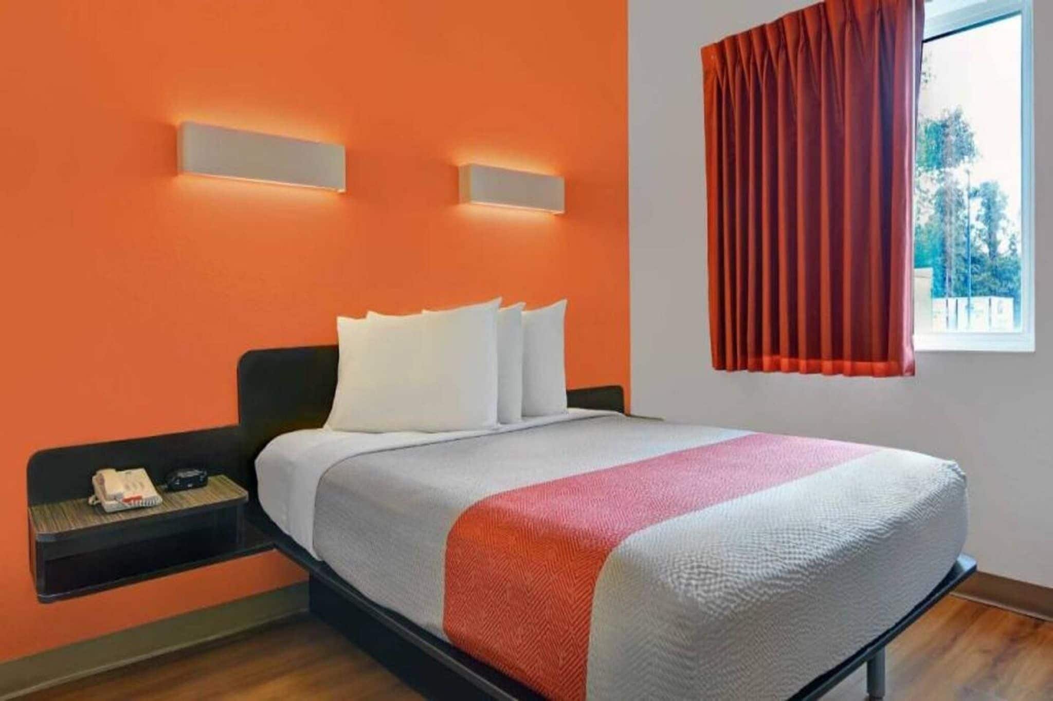 A hotel room with a bed, a nightstand, a window with curtains, and a wall-mounted light fixture.