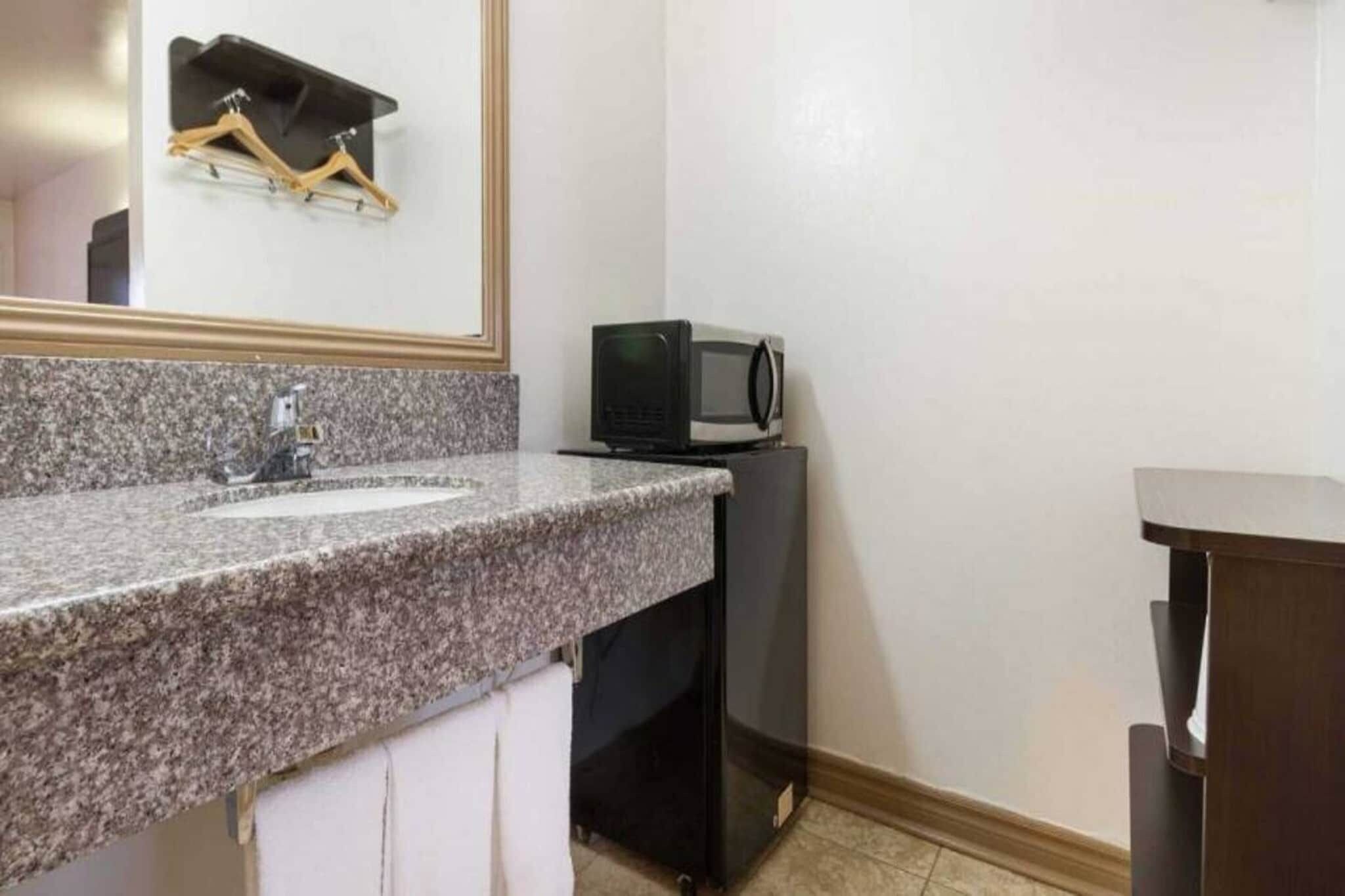 A bathroom with a granite countertop, a sink, a mirror, a towel rack, a microwave, and a cabinet.