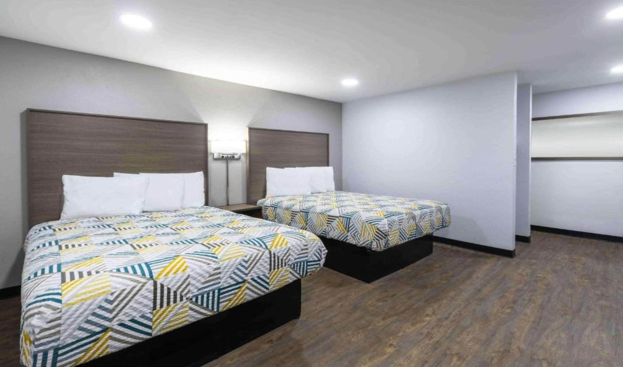 A hotel room with two beds, each with patterned bedding, a wooden headboard, a bedside table, and a wall-mounted lamp.