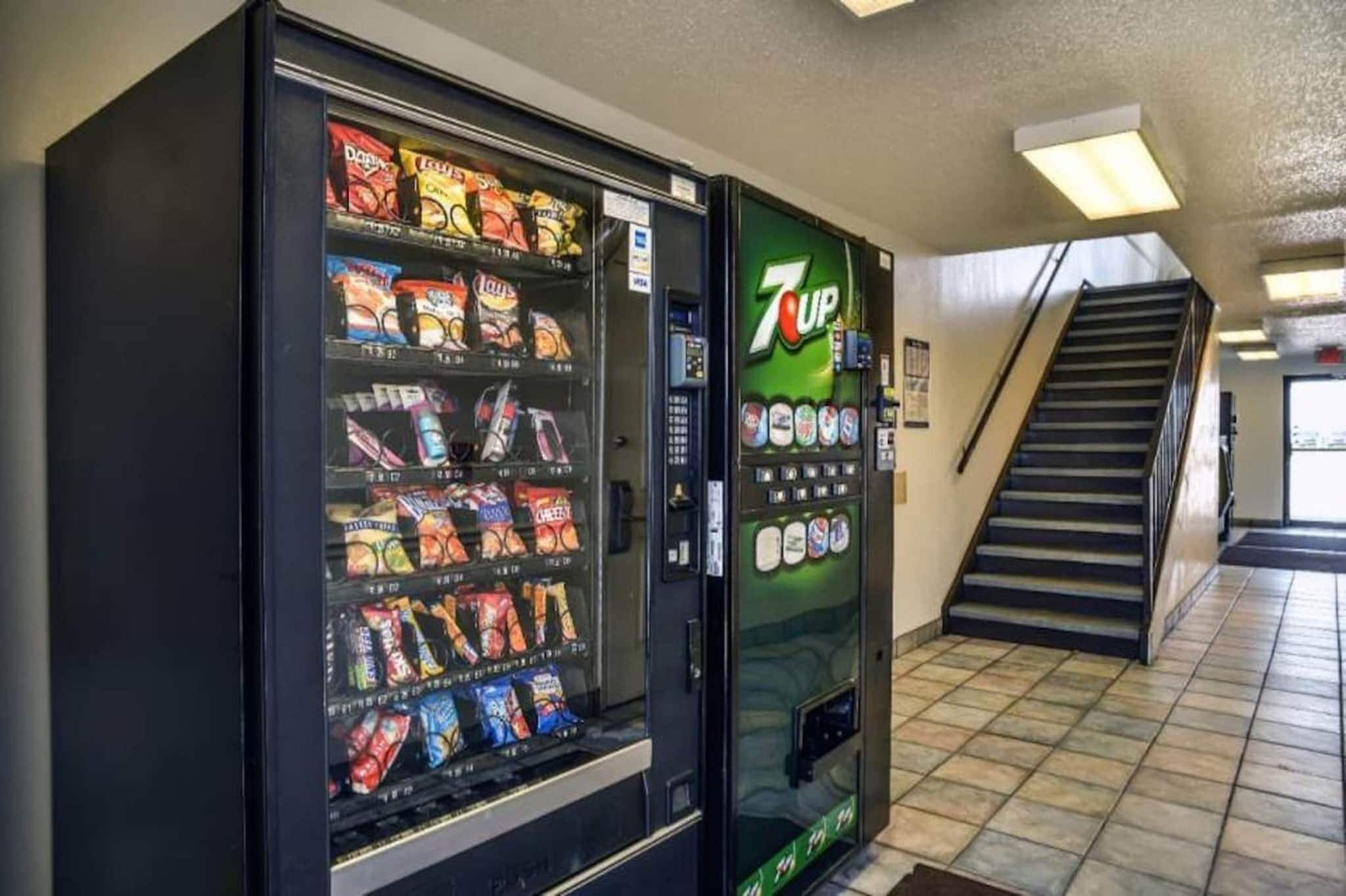 A vending machine with various snacks and a 7UP dispenser in a hallway with a staircase.