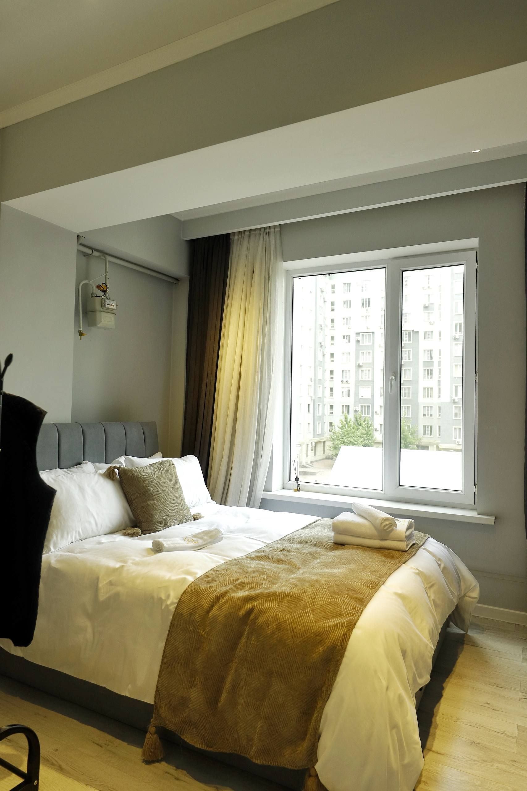 Ein modernes Schlafzimmer mit einem großen Bett, einem Fenster mit Blick auf Gebäude und einer Wandleuchte.