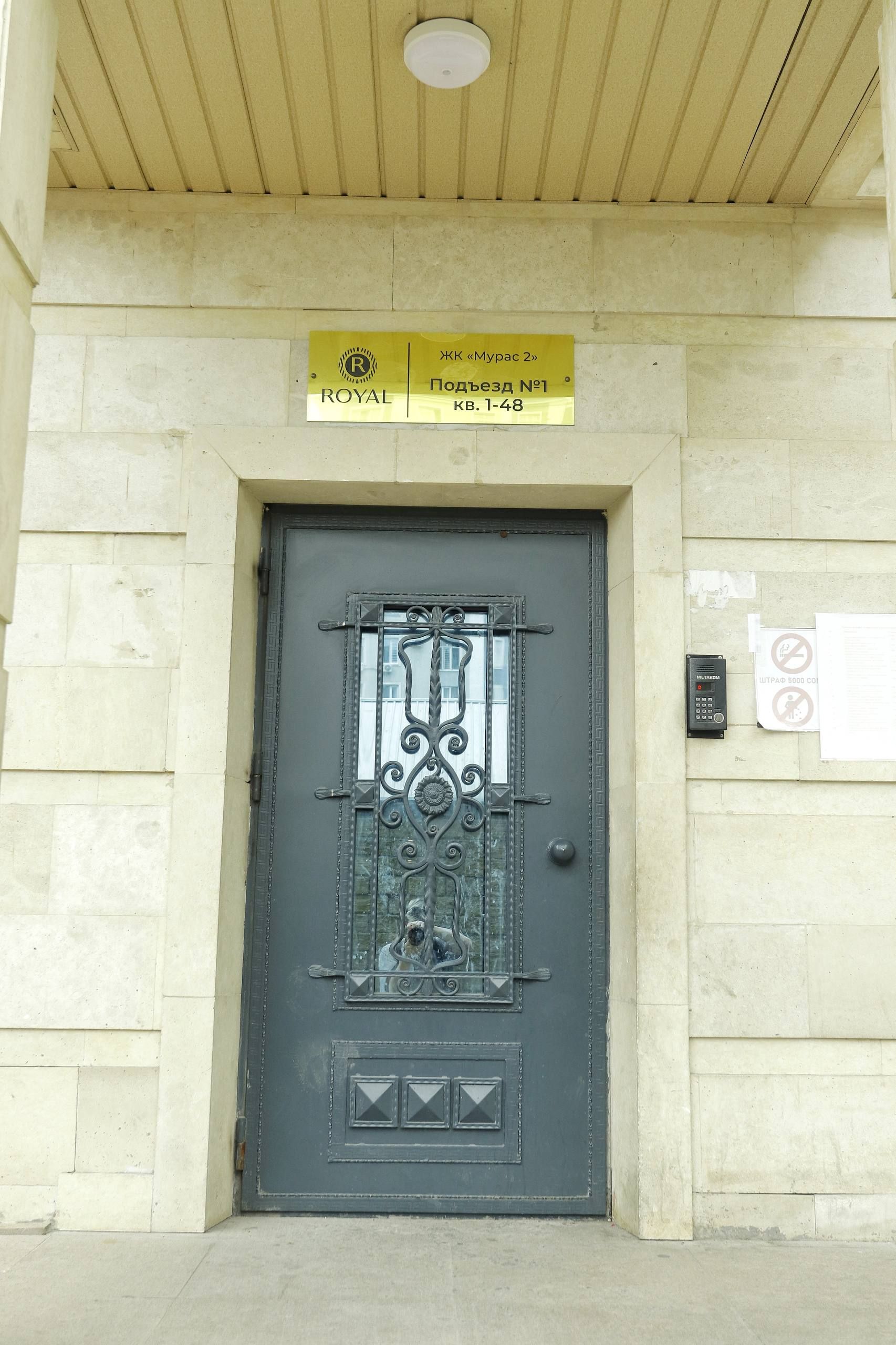 Une entrée d’immeuble avec une porte métallique, une enseigne au-dessus indiquant « ROYAL » et « Myras 2 », et un clavier de code sur le mur.