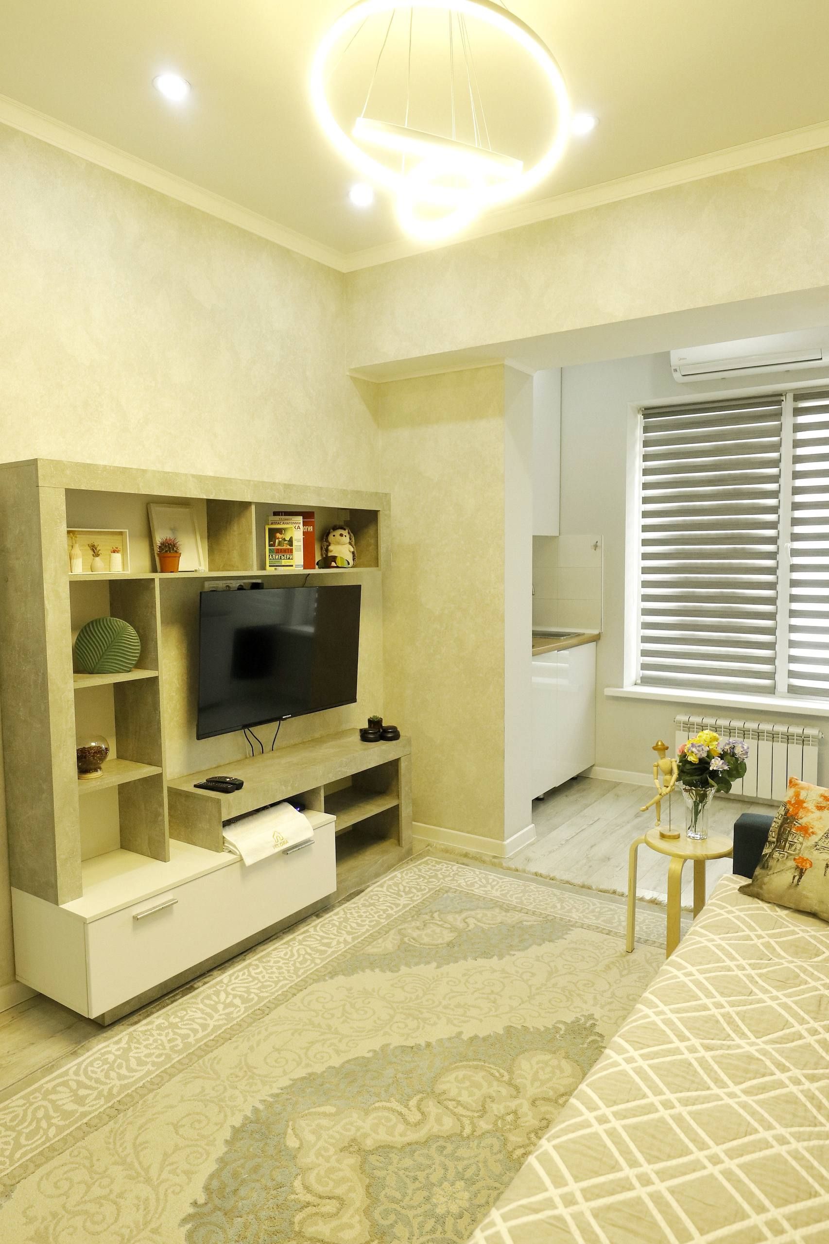 Ein modernes Wohnzimmer mit Flachbildfernseher, eingebauten Regalen, einem gemusterten Teppich und einem Fenster mit Jalousien.