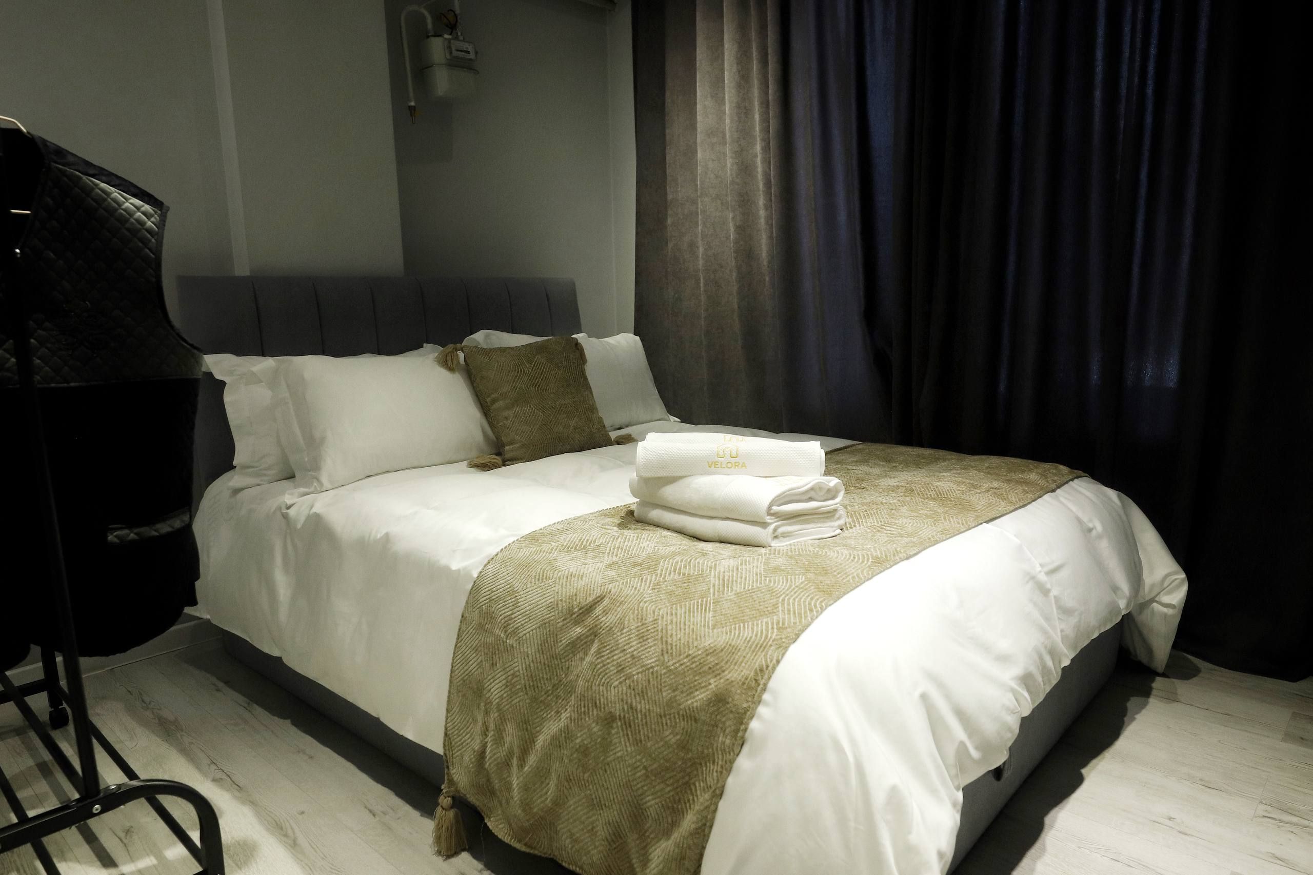 Un letto rifatto con lenzuola bianche e una coperta beige, una sedia nera e una tenda scura in una camera d'albergo.