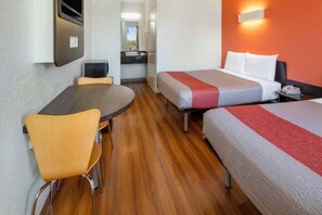 A hotel room with a wooden floor, a round dining table, two single beds with red and grey bedding, a small kitchenette, and a TV mounted on the wall.
