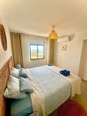 A hotel room with a bed, a window with curtains, a wall-mounted air conditioner, and a hanging light fixture. - Delightful 2-bedroom oceanview apartment with AC in Quarteira (Quarteira)