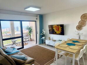 A modern living room with a sofa, a dining table with placemats, a TV mounted on the wall, and a view of the city through large windows. - Delightful 2-bedroom oceanview apartment with AC in Quarteira (Quarteira)