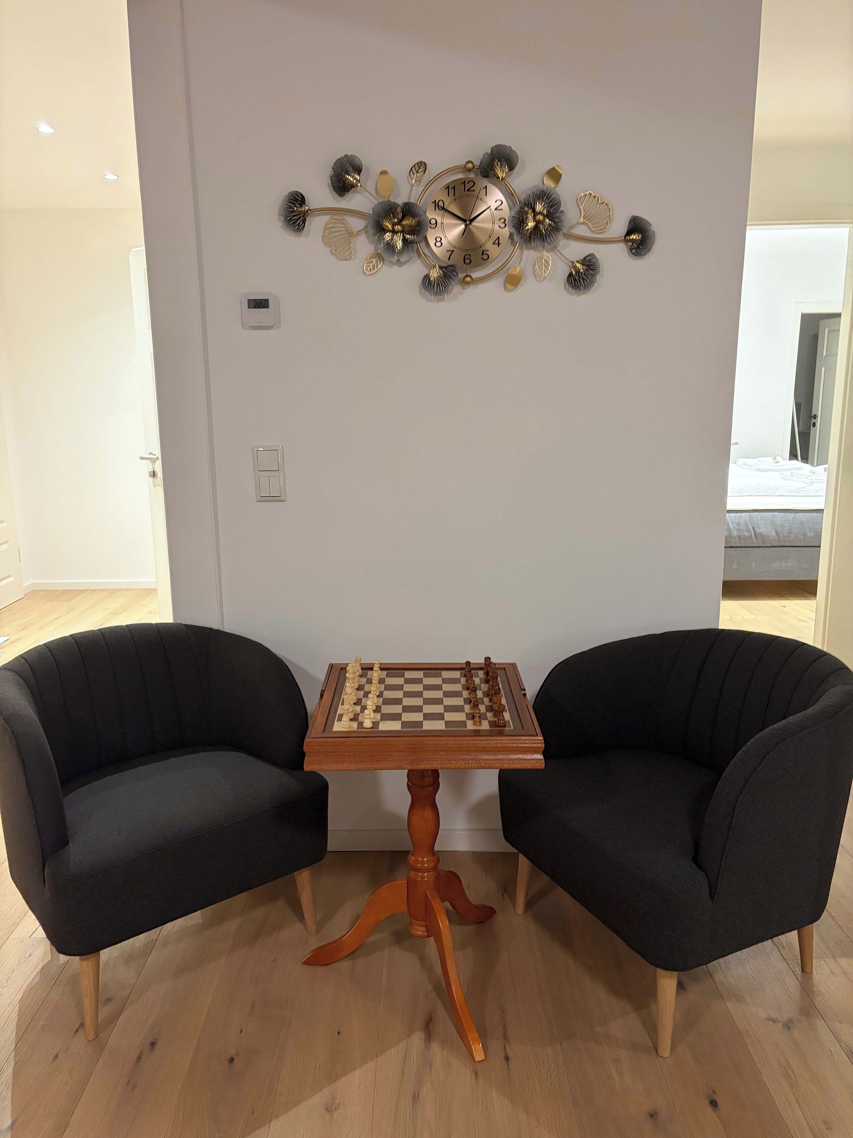 Una habitación con una mesa de ajedrez y dos sillones, un reloj de pared y una puerta.