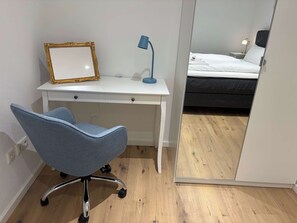 Une chambre avec un bureau blanc, une chaise bleue, un lit avec une literie blanche et un miroir qui reflète la pièce.