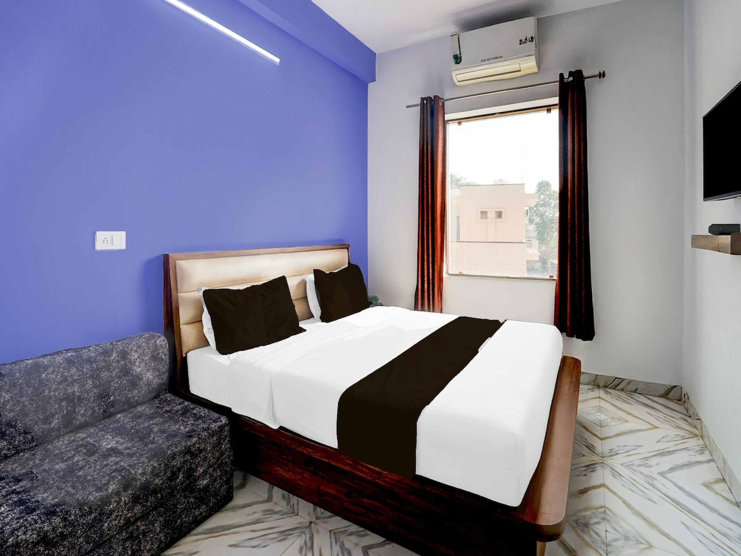 A hotel room with a bed, a sofa, a window with curtains, an air conditioning unit, and a television.