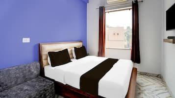 A hotel room with a bed, a sofa, a window with curtains, an air conditioning unit, and a television.