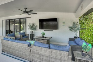 A covered outdoor seating area with wicker furniture, a ceiling fan, and a flat-screen TV mounted on the wall. - Outdoor Oasis! Luxe Waterfront Gem in Lehigh Acres (Lehigh Acres)
