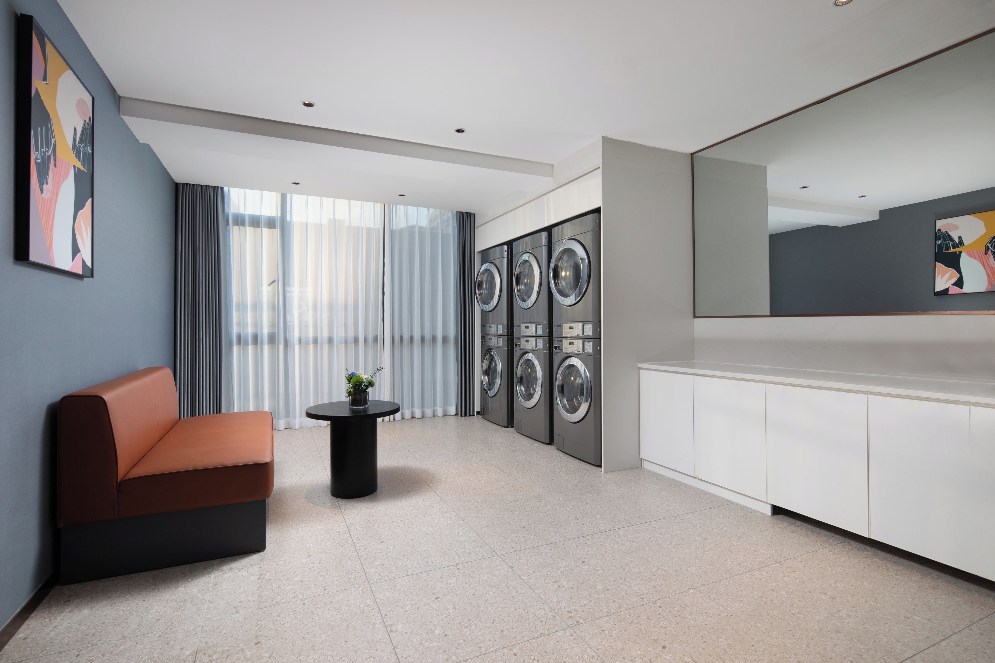 A modern hotel lobby with a sofa, a small table with a plant, a large window with curtains, and a row of washing machines.