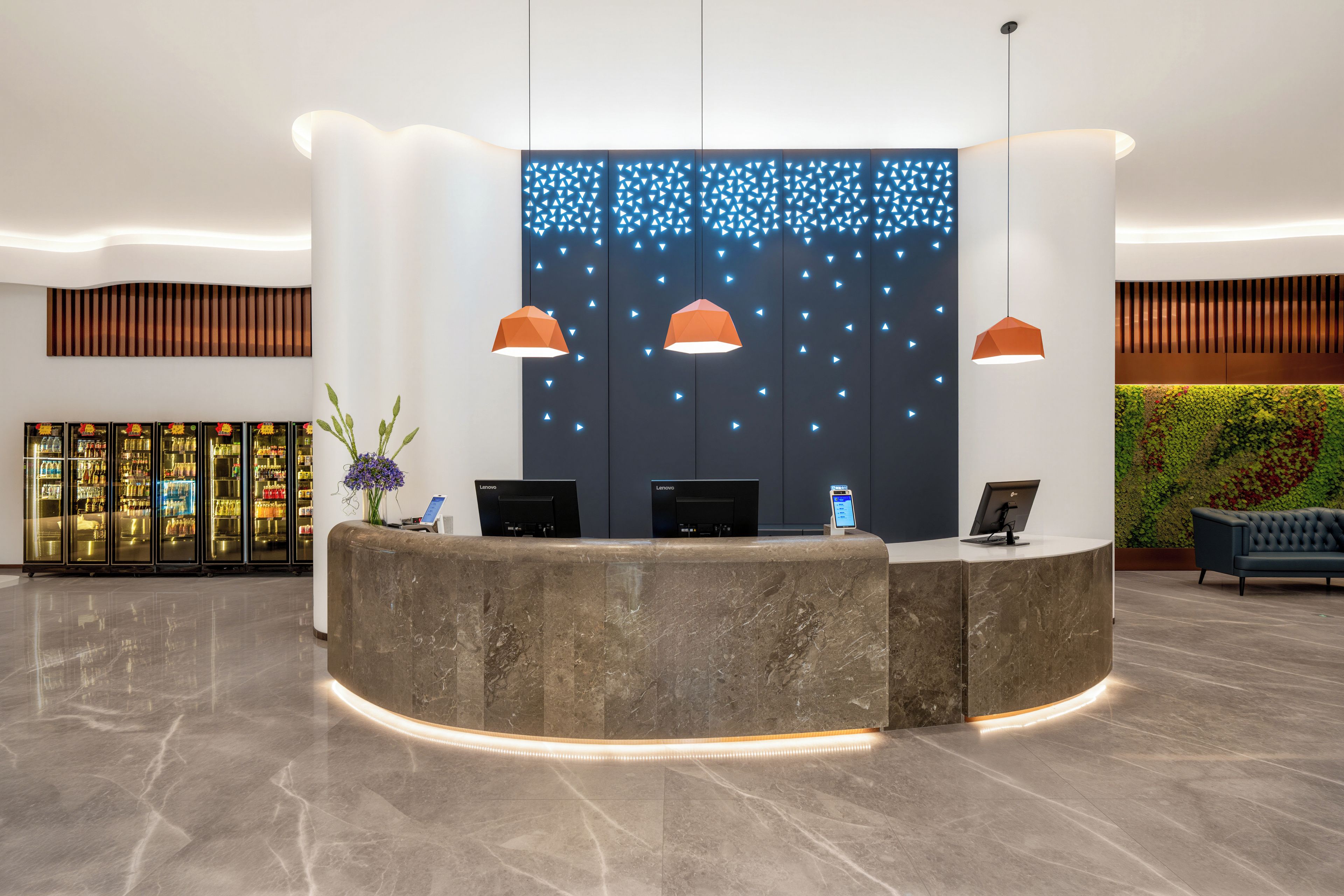 A reception desk with two monitors, a plant, and a wall with a geometric light pattern.
