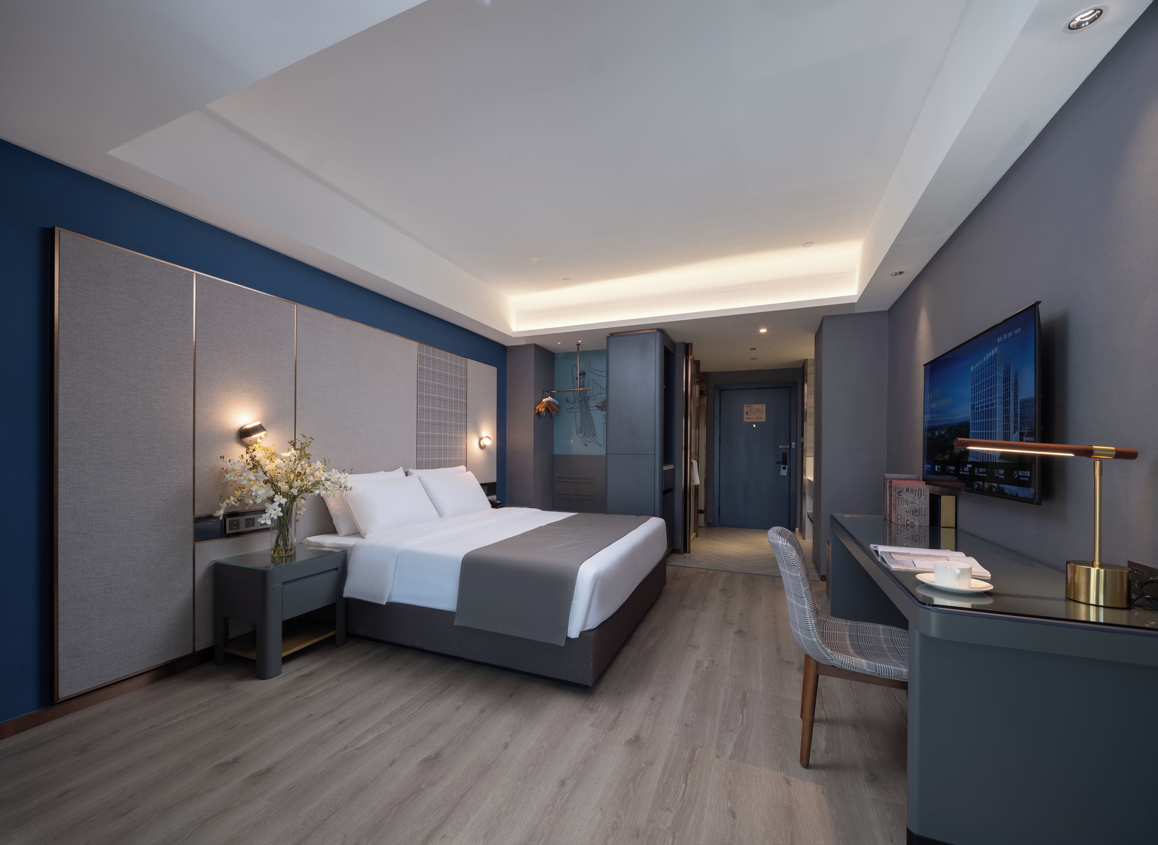 A modern hotel room with a large bed, bedside tables, a desk with a chair, a flat-screen TV mounted on the wall, and a door leading to the hallway.