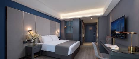 A modern hotel room with a large bed, bedside tables, a desk with a chair, a flat-screen TV mounted on the wall, and a door leading to the hallway.