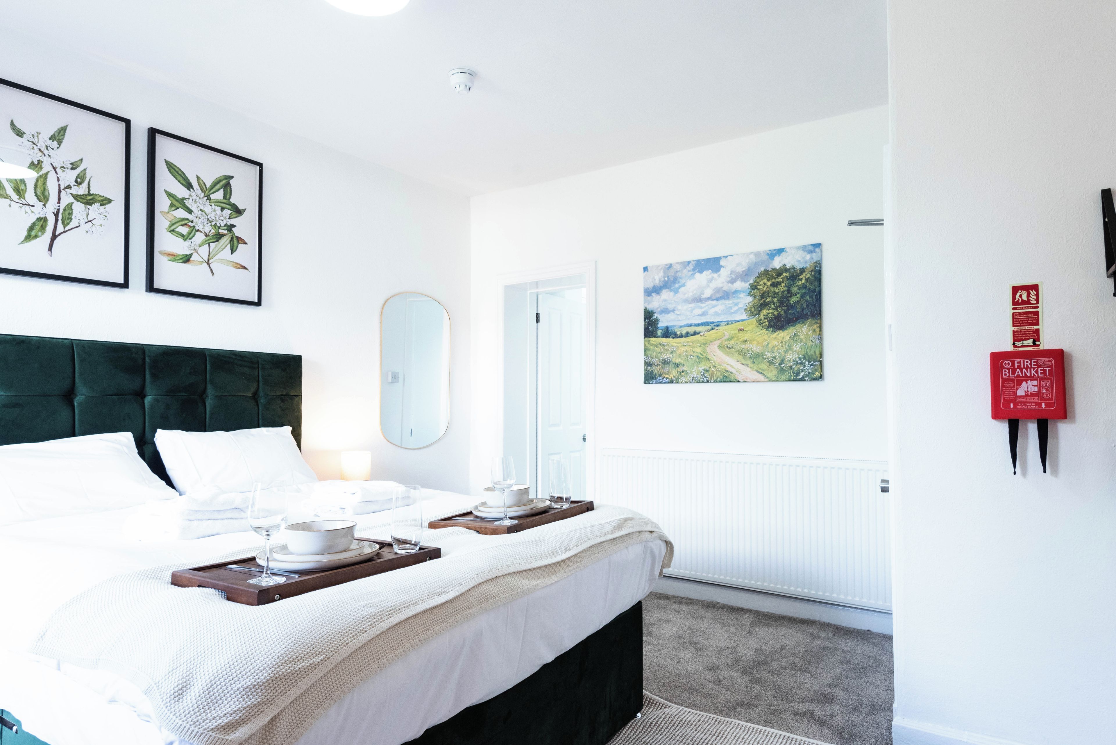 A hotel room with a bed, a bedside table with glasses and a bowl, a mirror, framed botanical prints, a painting, and a fire blanket on the wall.