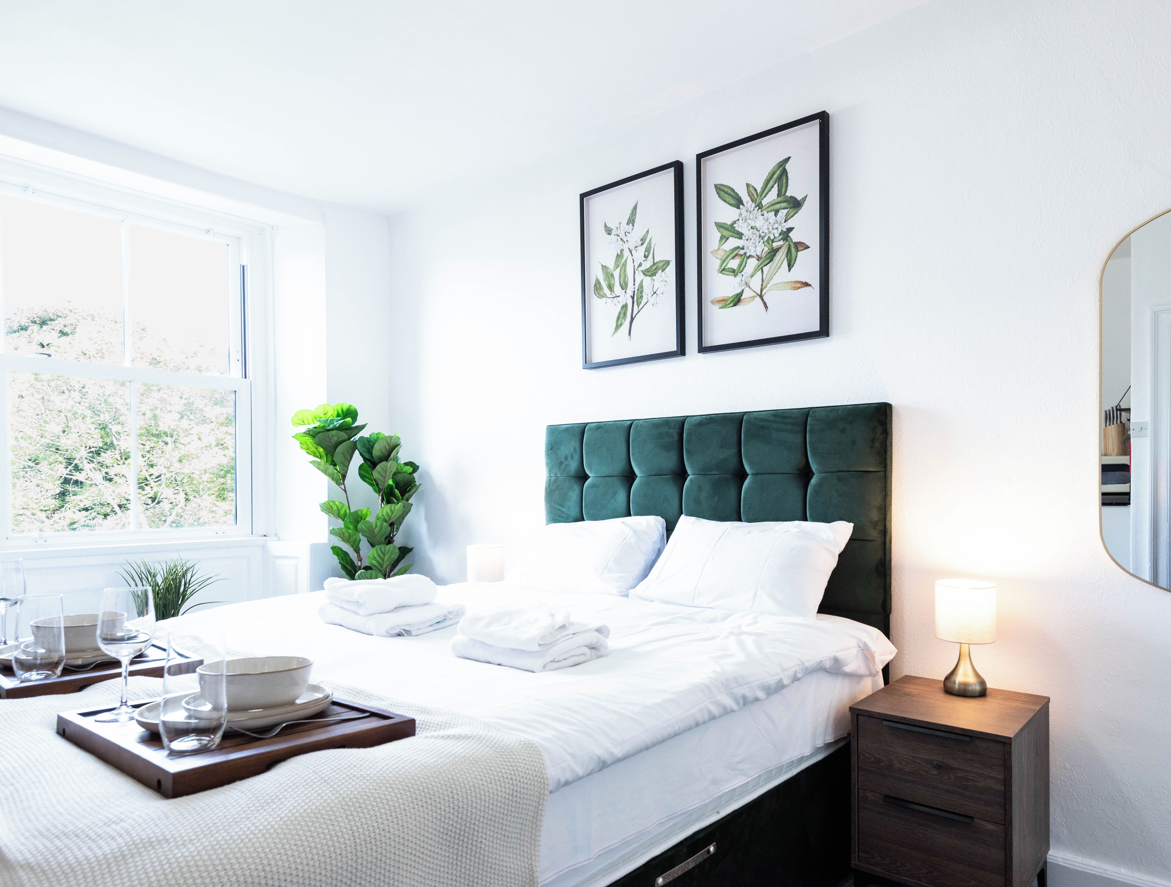 A neatly made bed with white linens, a green upholstered headboard, and a bedside table with a lamp. There are framed botanical prints on the wall above the bed.