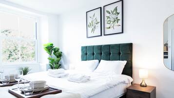 A neatly made bed with white linens, a green upholstered headboard, and a bedside table with a lamp. There are framed botanical prints on the wall above the bed.