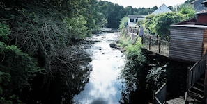 Une rivière traverse une zone boisée, et on y aperçoit un pont en bois ainsi que des bâtiments situés sur la rive droite.