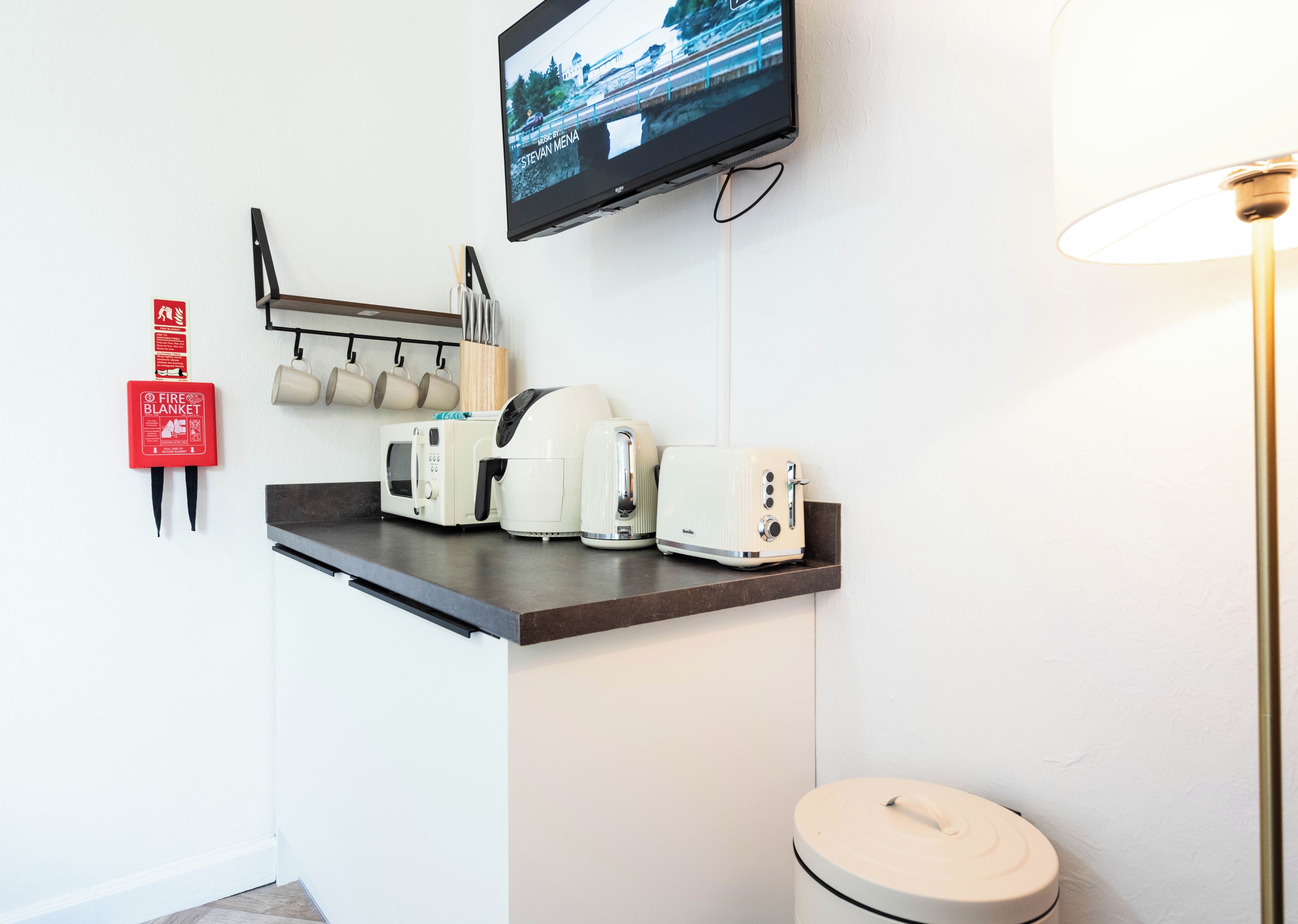 A kitchenette with a microwave, toaster, and coffee maker on a countertop, a fire blanket on the wall, and a TV mounted above.