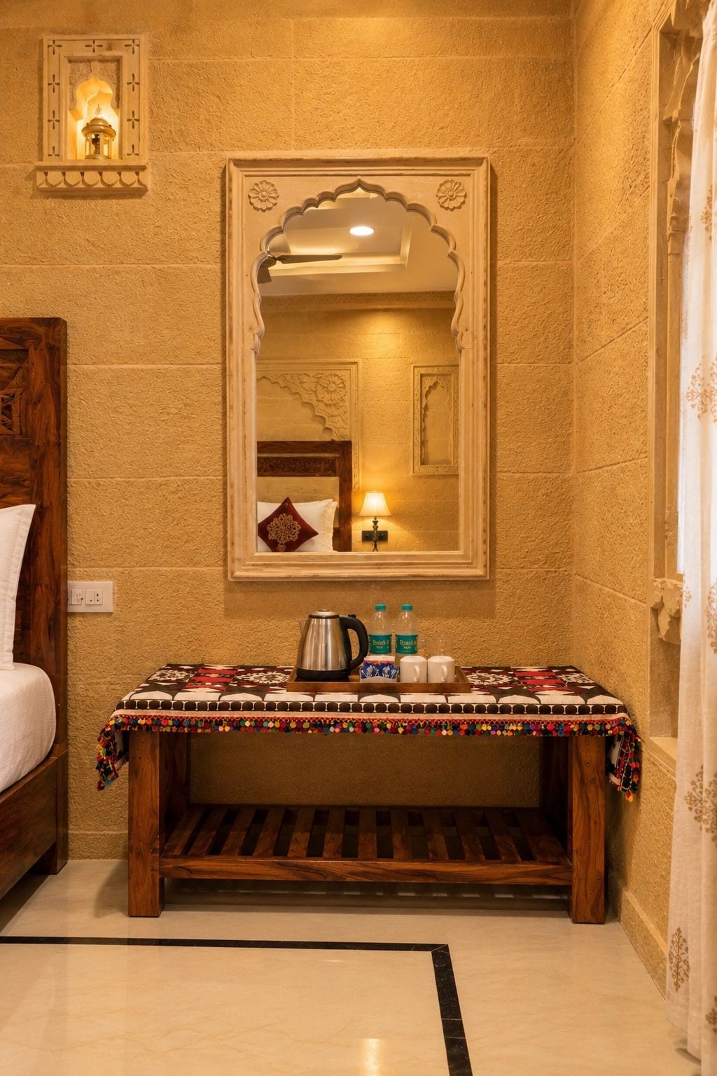A hotel room with a bed, a wooden bench, a table with a tray, a mirror, and a wall-mounted light fixture.