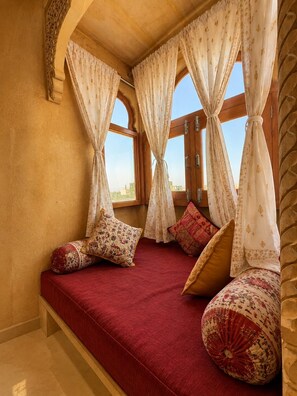 A room with a red cushioned bench, patterned pillows, and a view through a window with curtains. - HOTEL ROYAL LAKHINA JAISALMER (Jaisalmer)