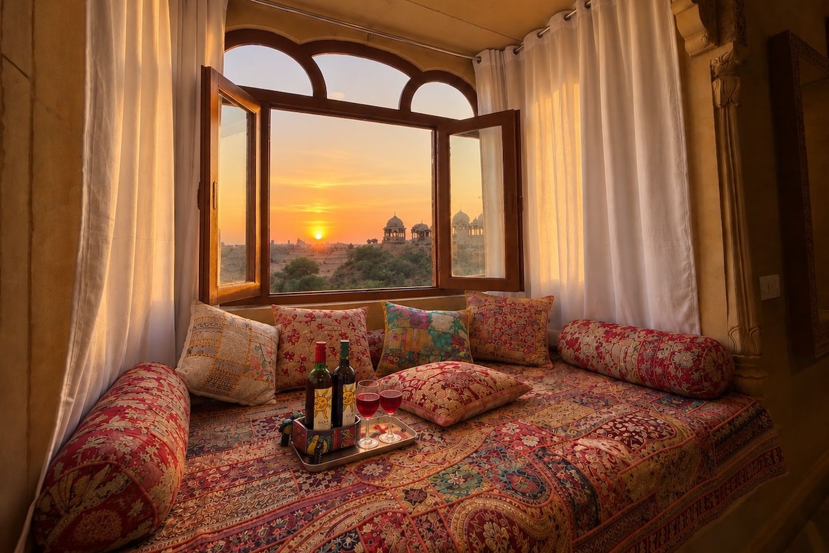 A room with a large window, a patterned bedspread, and a tray with bottles and glasses.