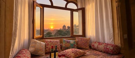 A room with a large window, a patterned bedspread, and a tray with bottles and glasses.