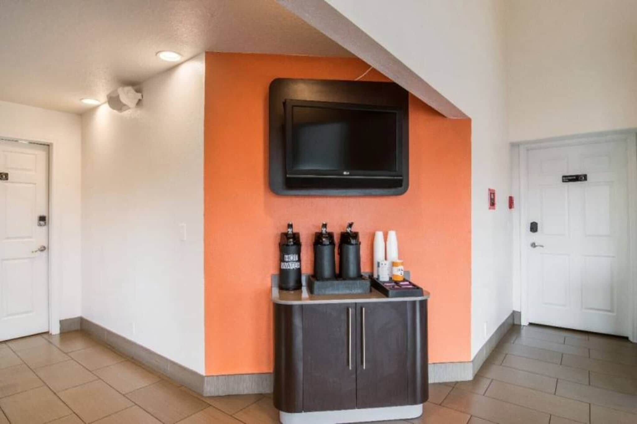 Un hall d’hôtel avec un téléviseur à écran plat fixé sur un mur orange, un comptoir de réception doté d’un meuble sombre et des portes blanches.