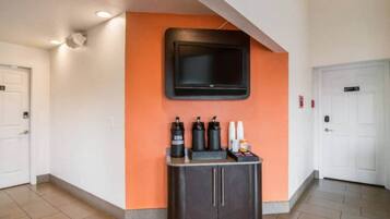 Un hall d’hôtel avec un téléviseur à écran plat fixé sur un mur orange, un comptoir de réception doté d’un meuble sombre et des portes blanches.