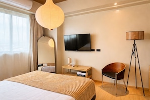 Una habitación de hotel con una cama grande, un televisor de pantalla plana, una mesita de noche con una lámpara, un lámpara de pie y un espejo.