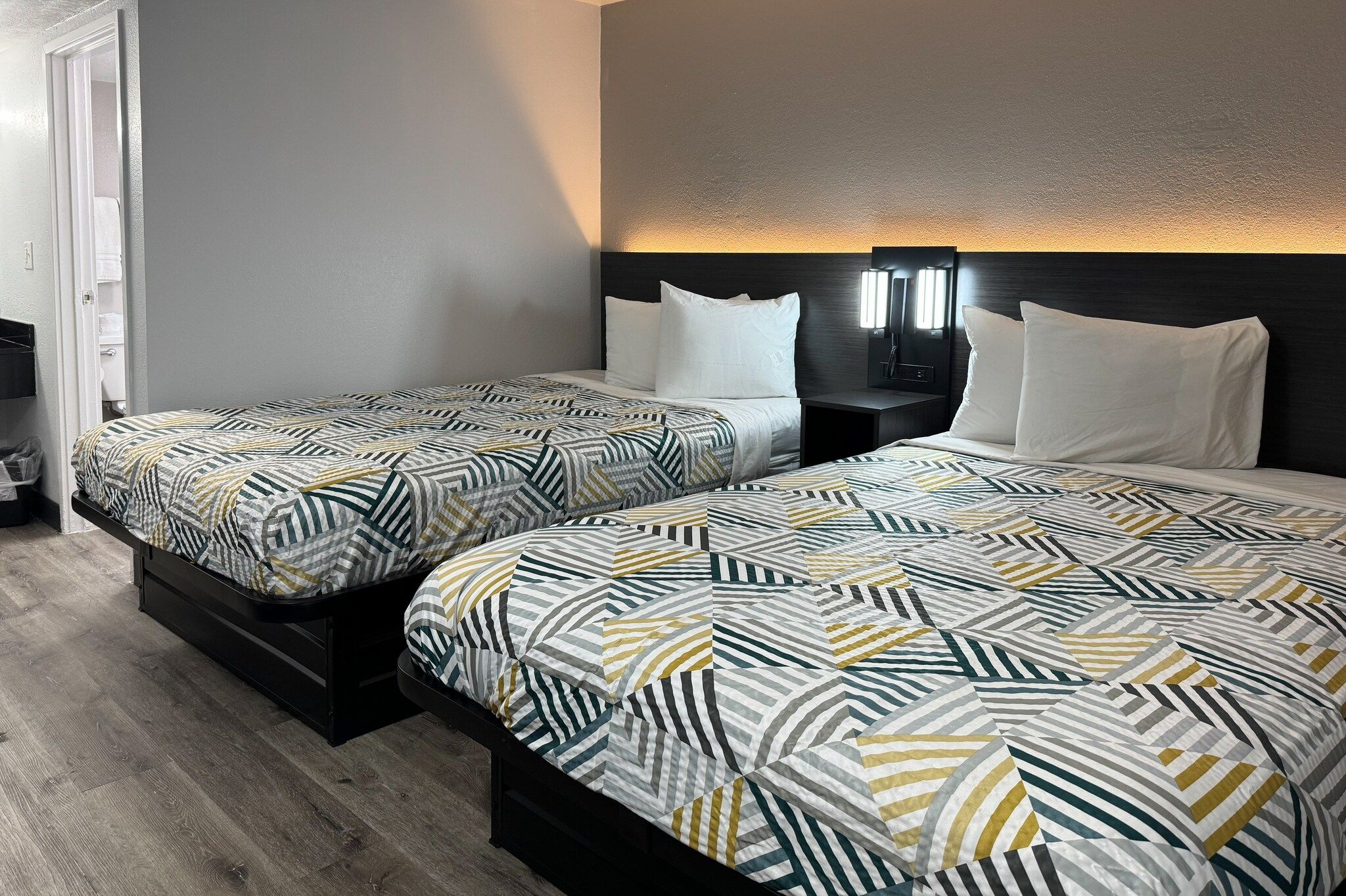 A hotel room with two beds, each with patterned bedding, a bedside table with a lamp, and a wall-mounted light fixture.