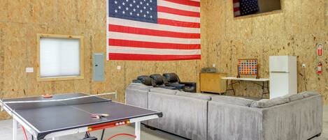 Una habitación con mesa de ping pong, sofá gris, televisor y bandera estadounidense.