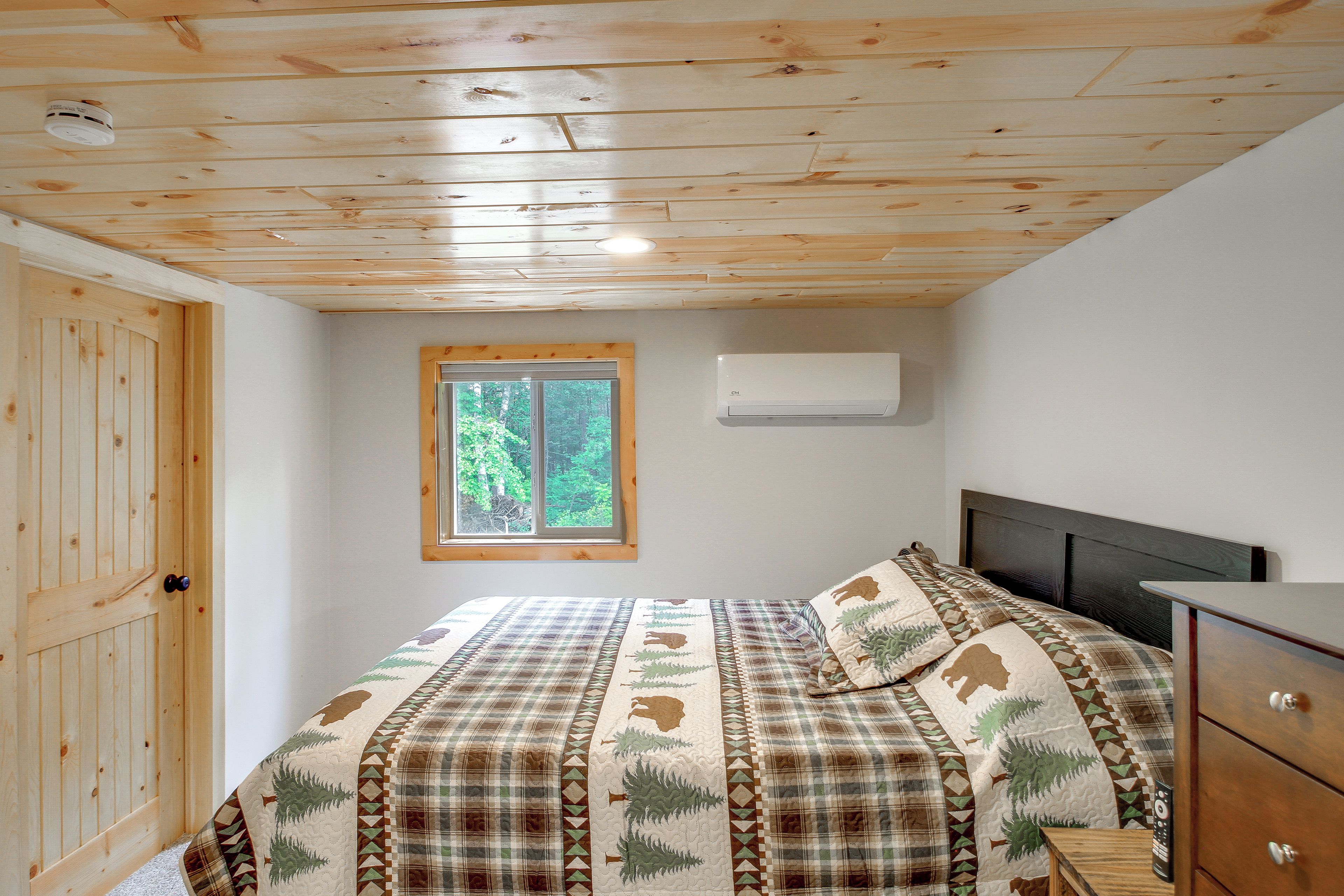 A bedroom with a wooden ceiling, a window with a view of trees, a bed with a patterned bedspread, and a bedside table with a remote control.